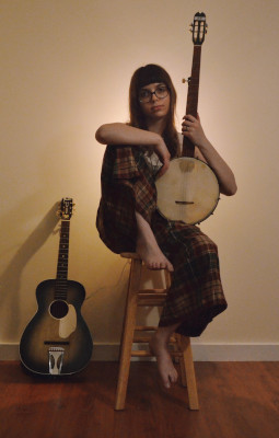 Lauren sitting on a bar stool holding a banjo upright, with a guitar standing against a wall on her right